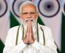 An Indian political leader standing with folded hands in front of national flags during an official event