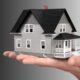 A hand holding a small house model with a bank loan approval symbol, representing financial support for home reconstruction